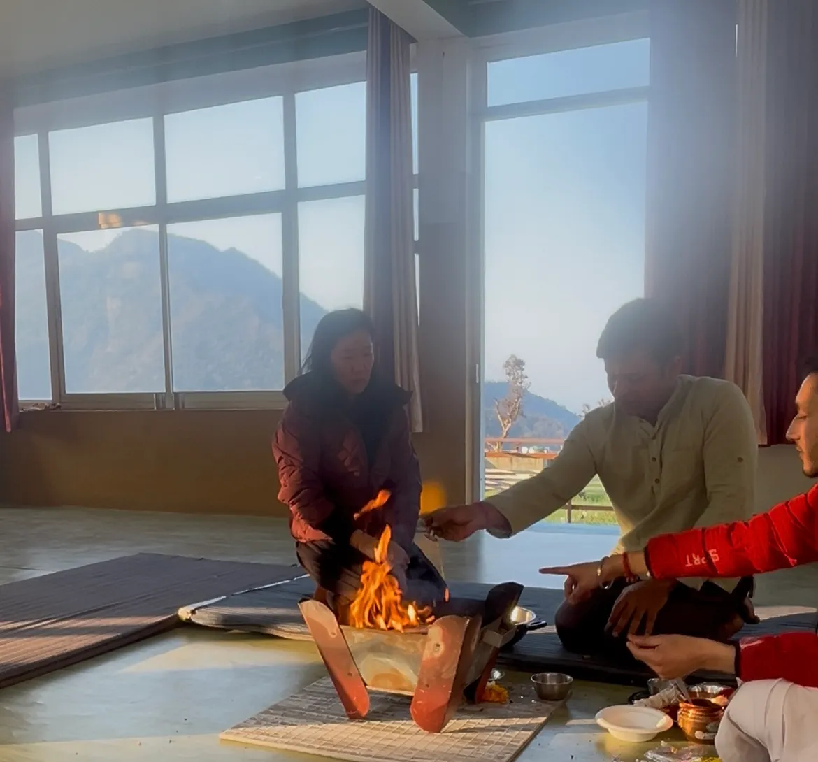 People sit around a small havan fire on the floor of a yoga hall, one placing an offering into the flames, hills through the windows