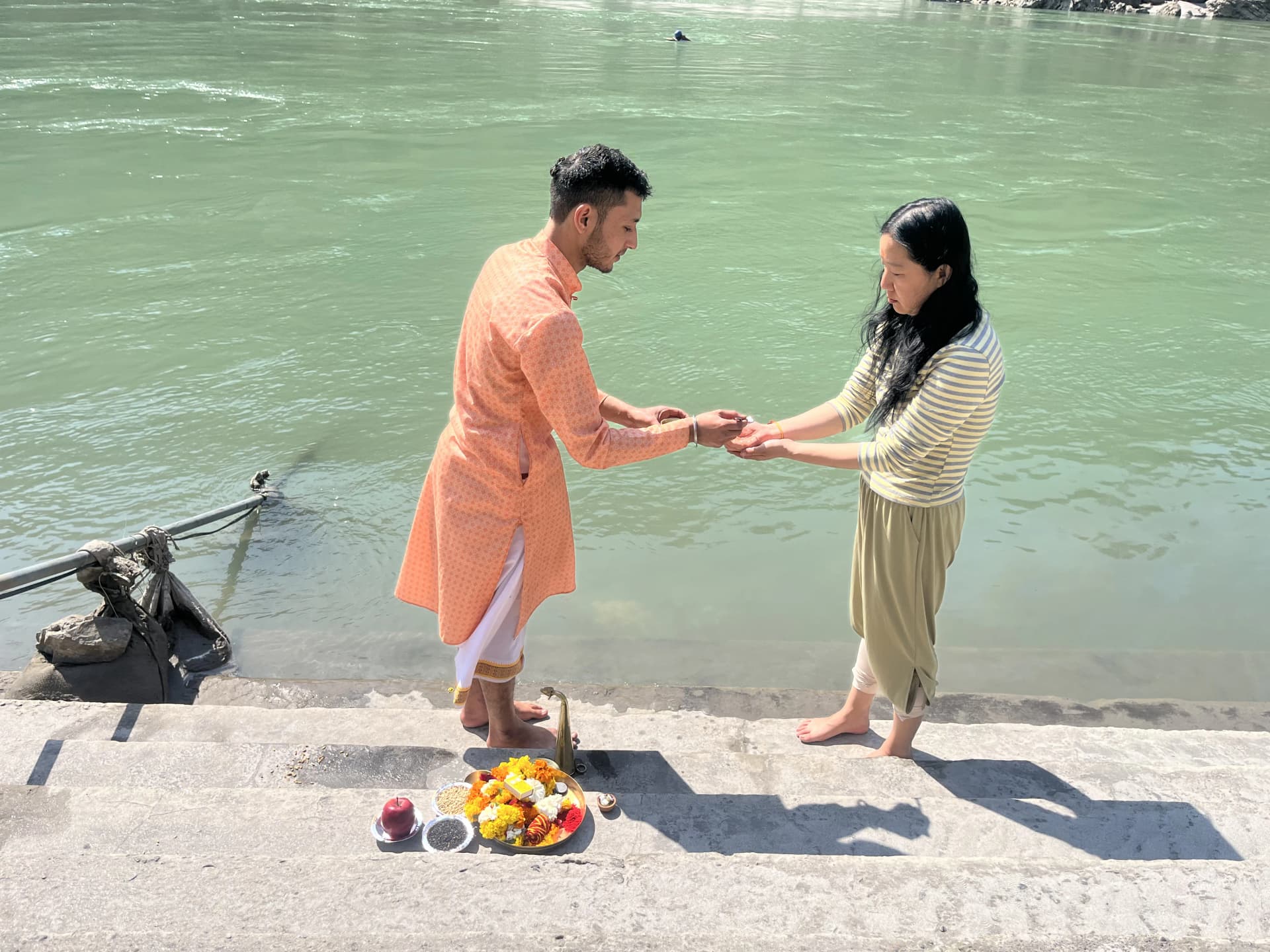 A man in an orange kurta and a woman stand facing each other on the Ganga ghat steps in Rishikesh, a plate of flowers and fruit beside them