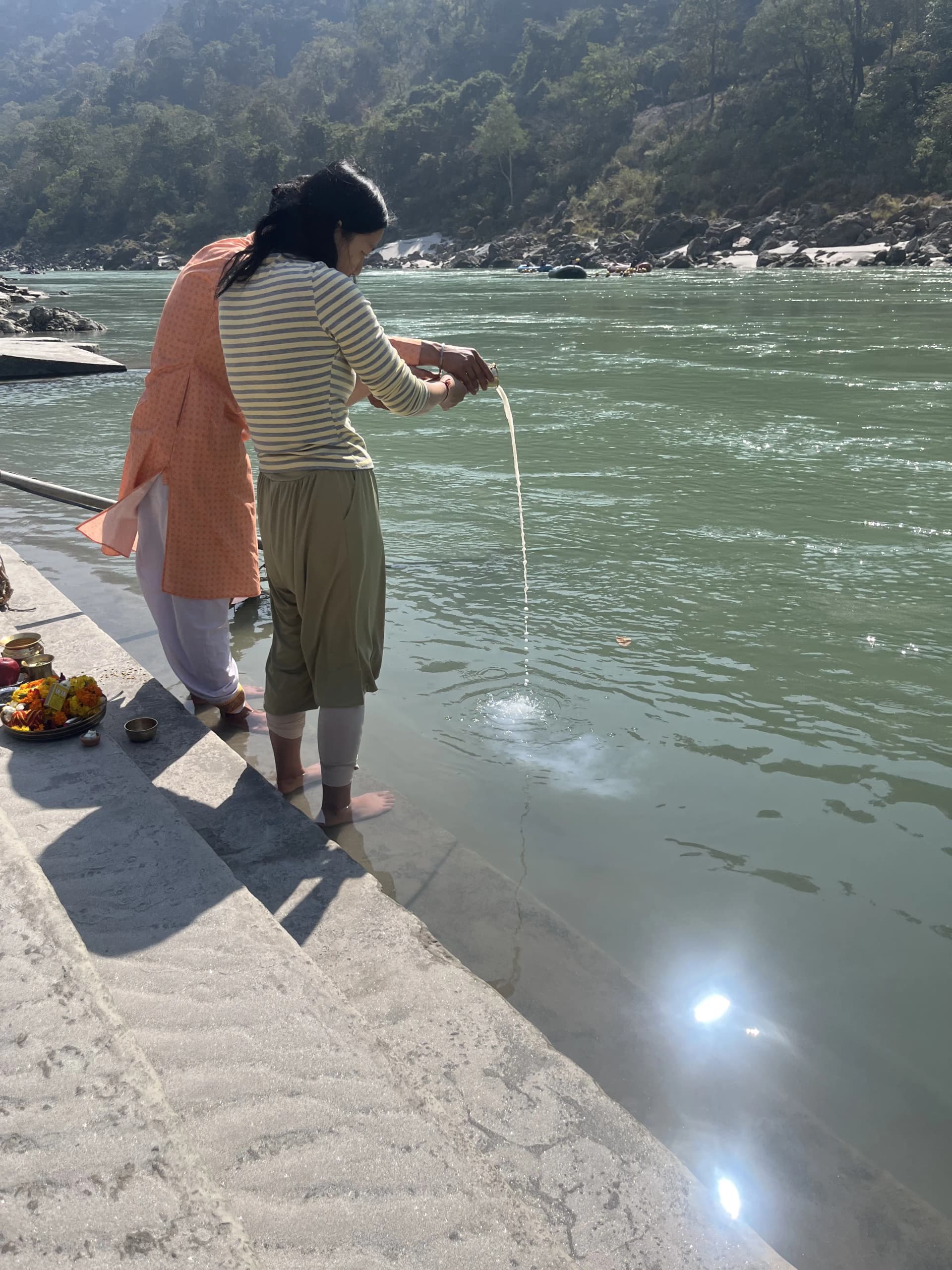 A woman pours water into the Ganga from the ghat steps in Rishikesh, a plate of flower offerings behind her