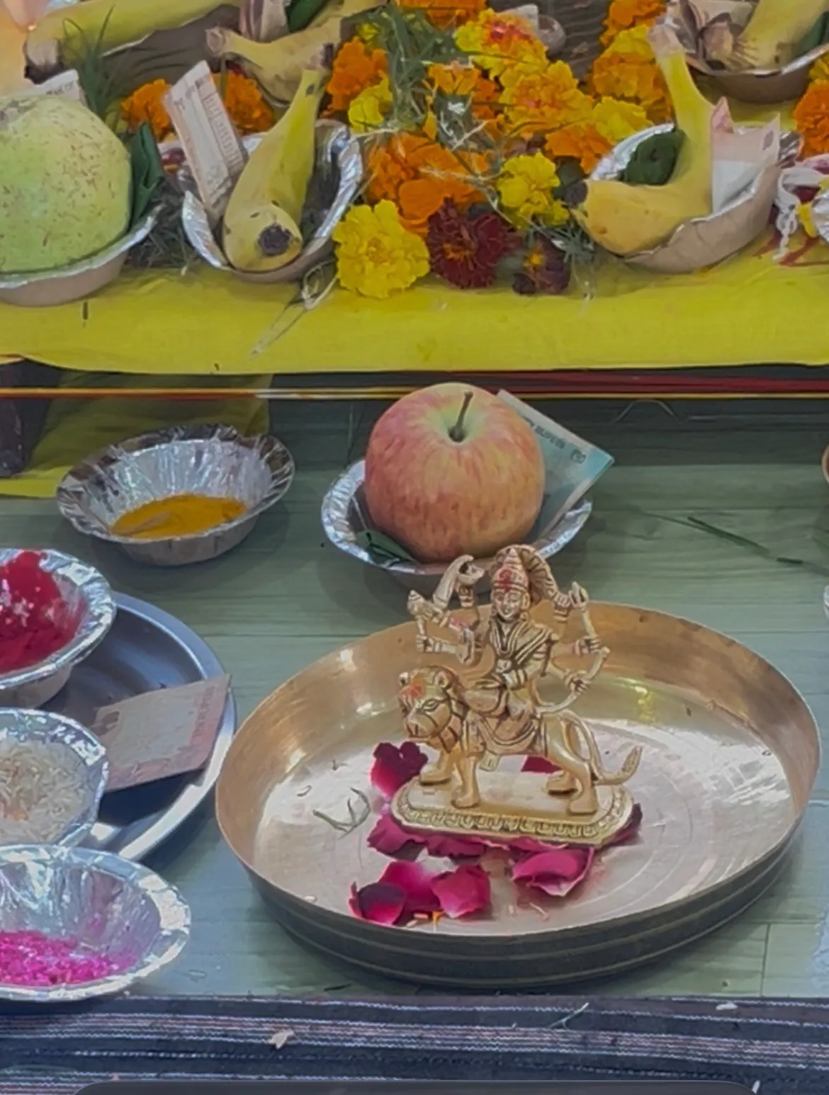 A small brass figure of Durga on a lion, on a brass plate with rose petals, fruit and marigolds behind