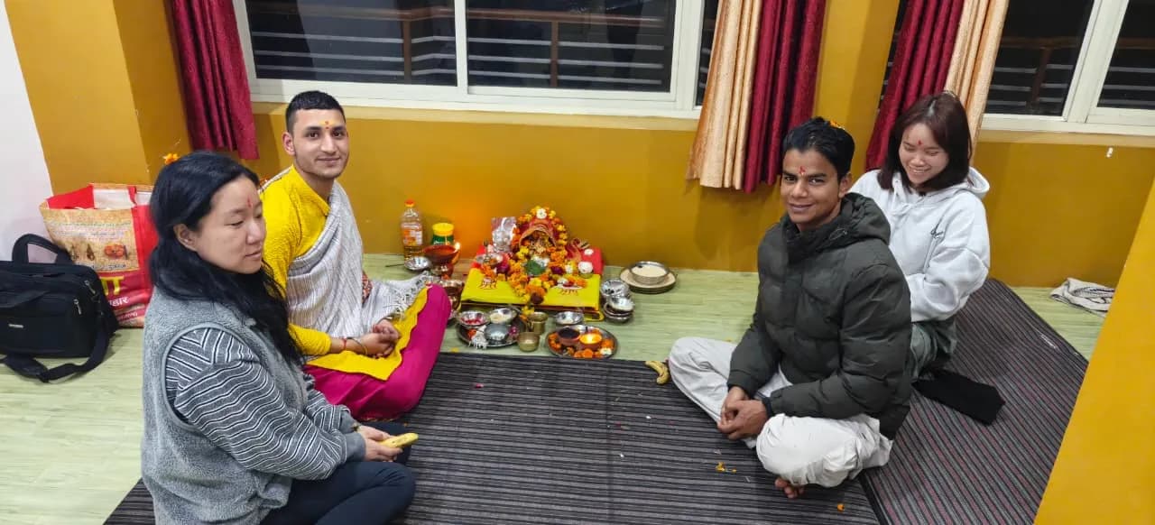 Four people sit on the floor around a puja altar decorated with marigolds and oil lamps in a yellow-walled room
