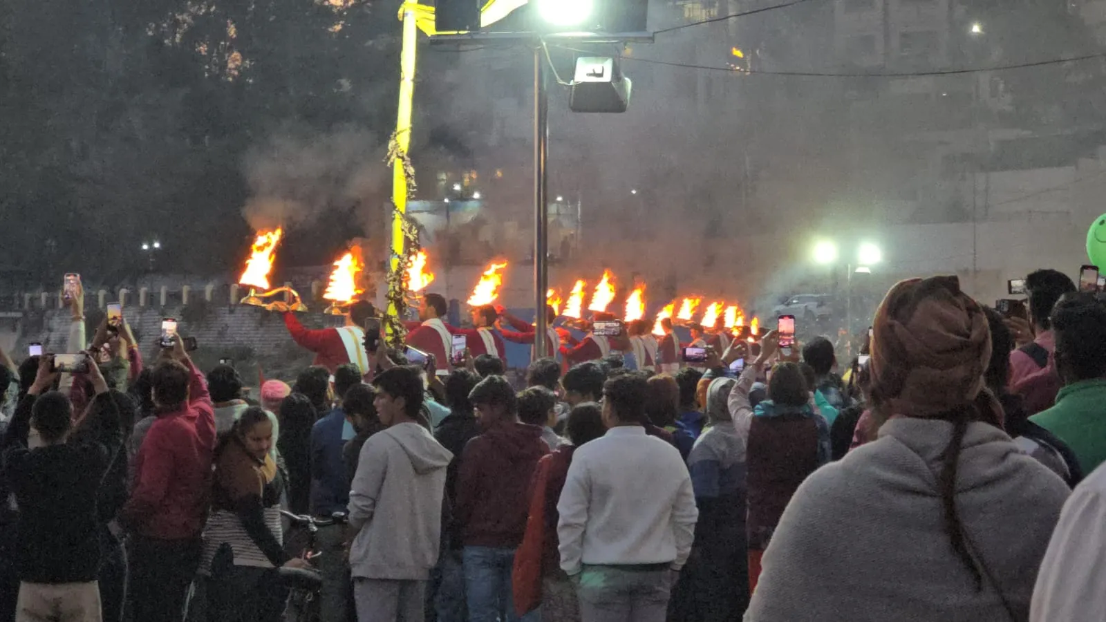 Priests raise flaming lamps at an evening Ganga aarti while a crowd watches and films