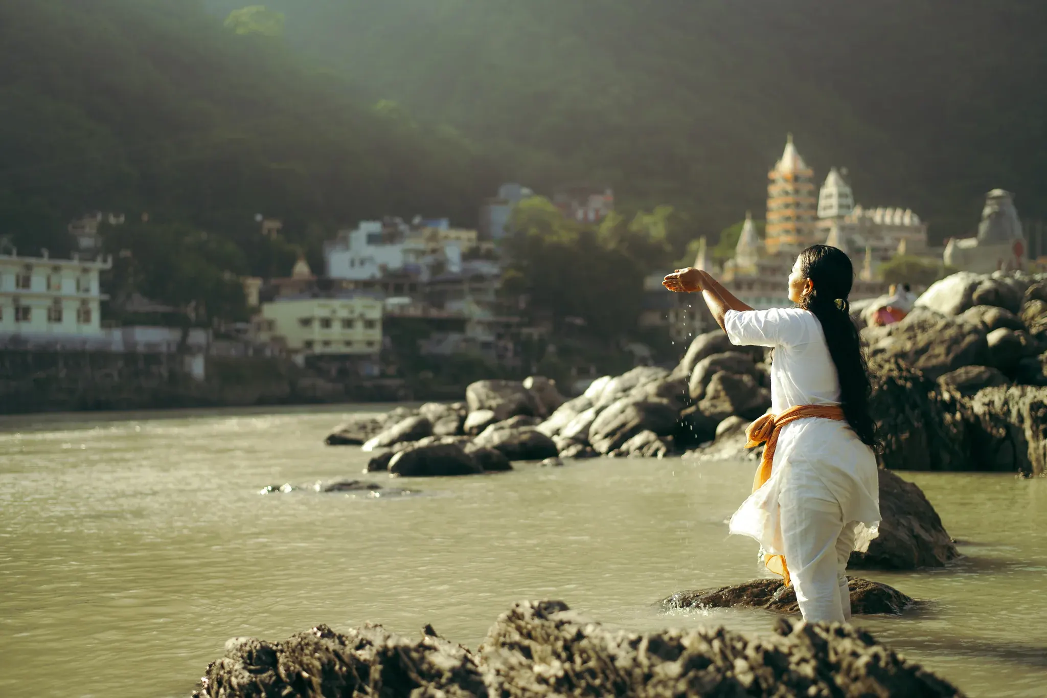Woman in white offers Ganga water from cupped hands toward the sun in Rishikesh, Trimbakeshwar temple behind