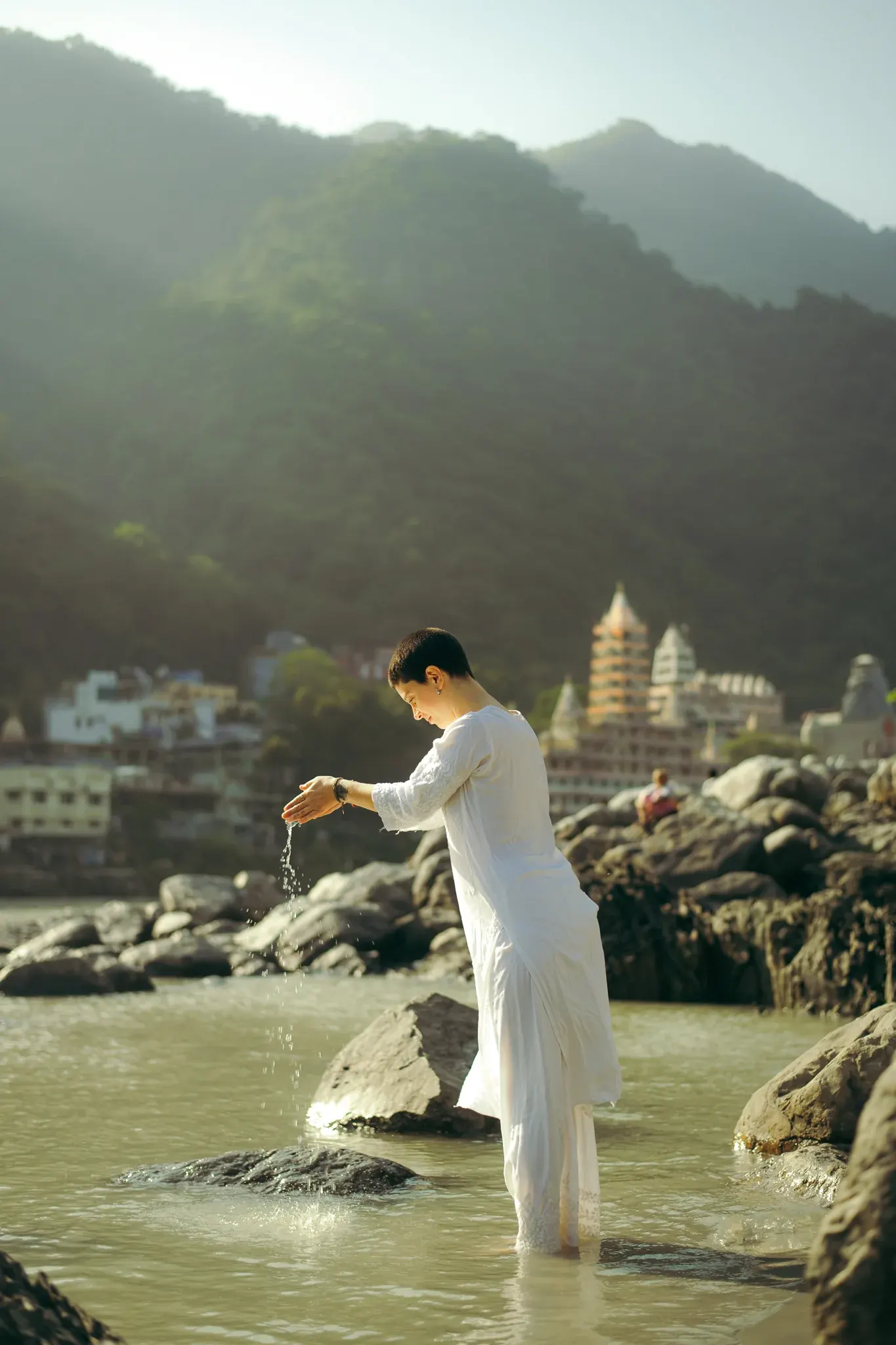Smiling woman in white stands in the Ganga releasing water from cupped hands, temple behind, Rishikesh