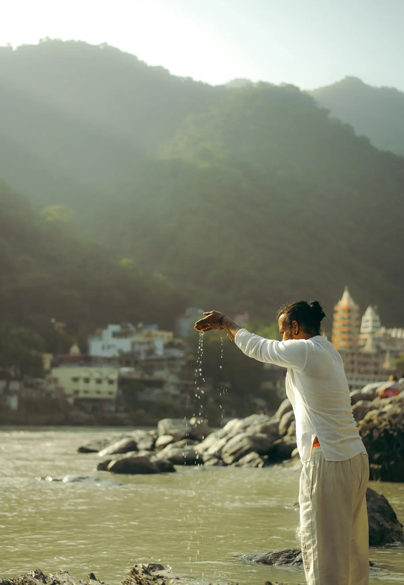 Man in white pours Ganga water from his raised hand as an offering, temple and hills of Rishikesh behind