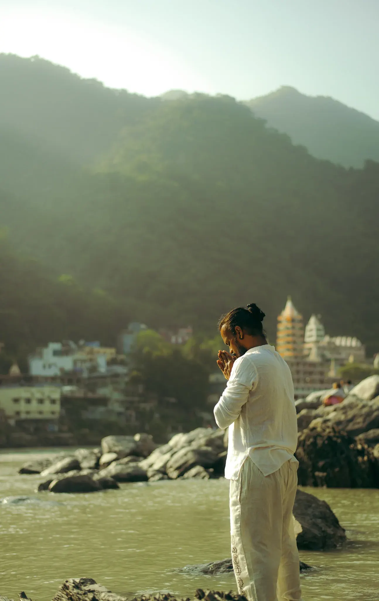 Man in white stands in the Ganga in Rishikesh with palms joined in prayer, head bowed, temple and hills behind