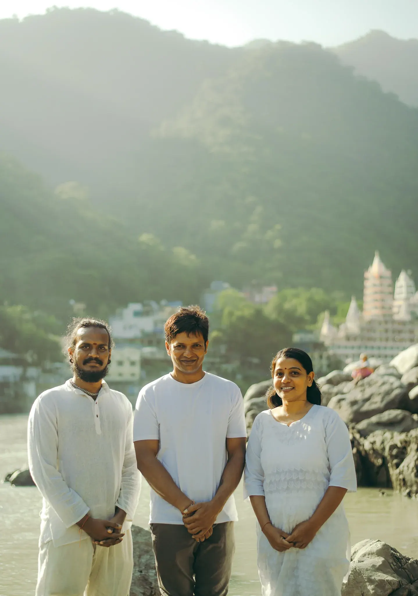 Yoga teacher stands smiling between two companions in white on the Ganga riverbank in Rishikesh