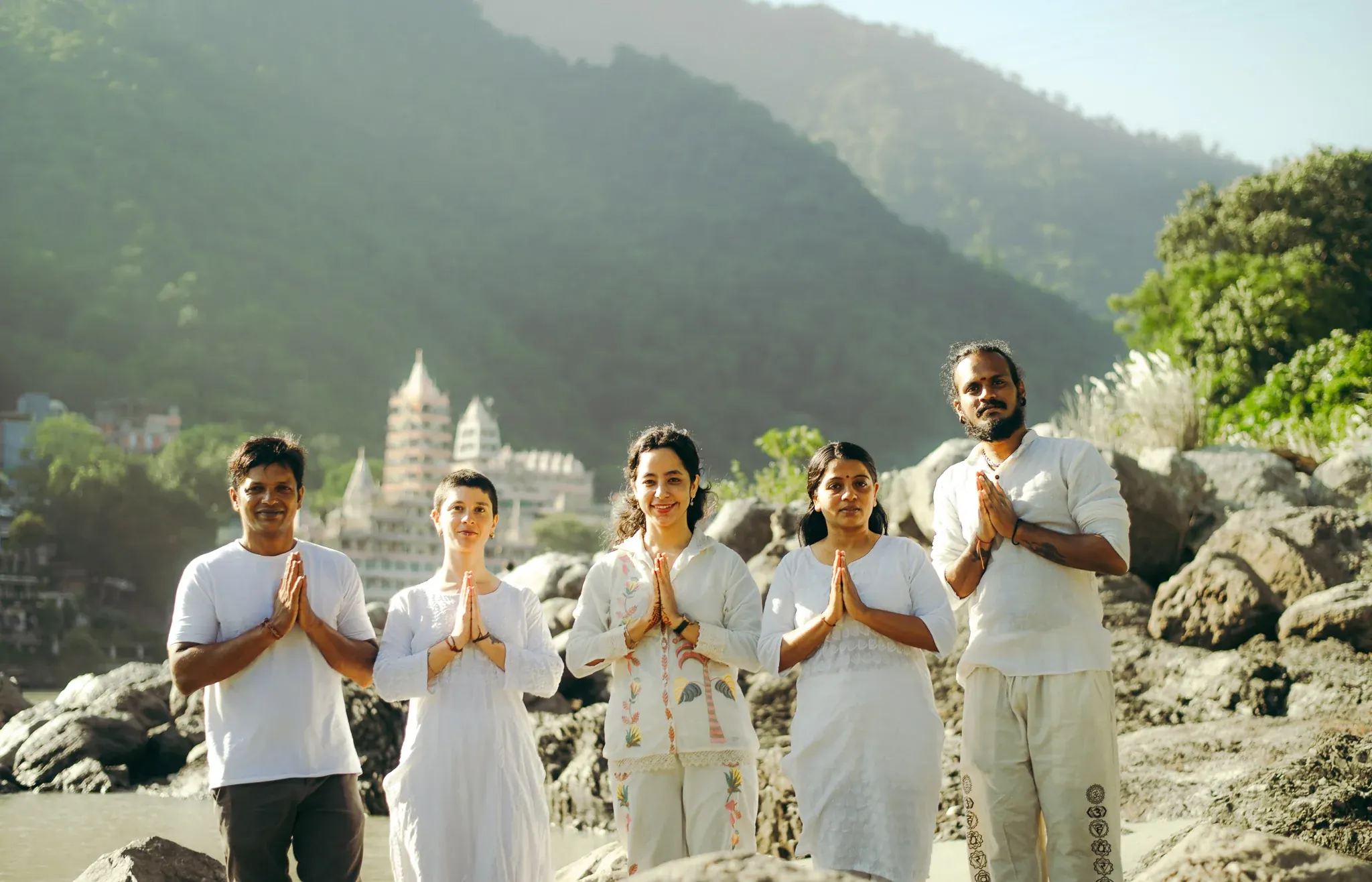 Teacher and four students in white stand with palms joined in namaste on the Ganga riverbank in Rishikesh