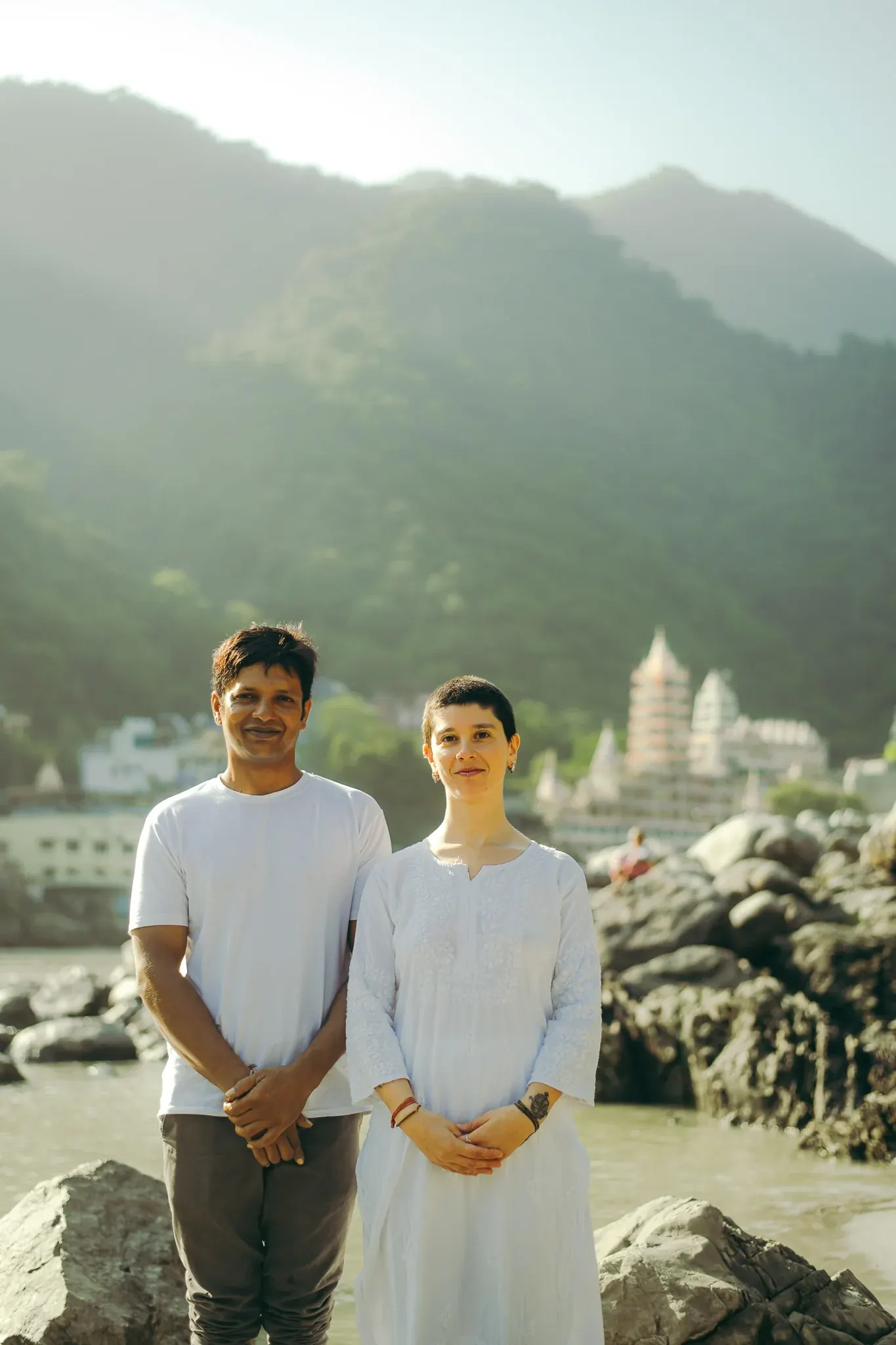 Yoga teacher and a student in white stand smiling together on the Ganga riverbank in Rishikesh