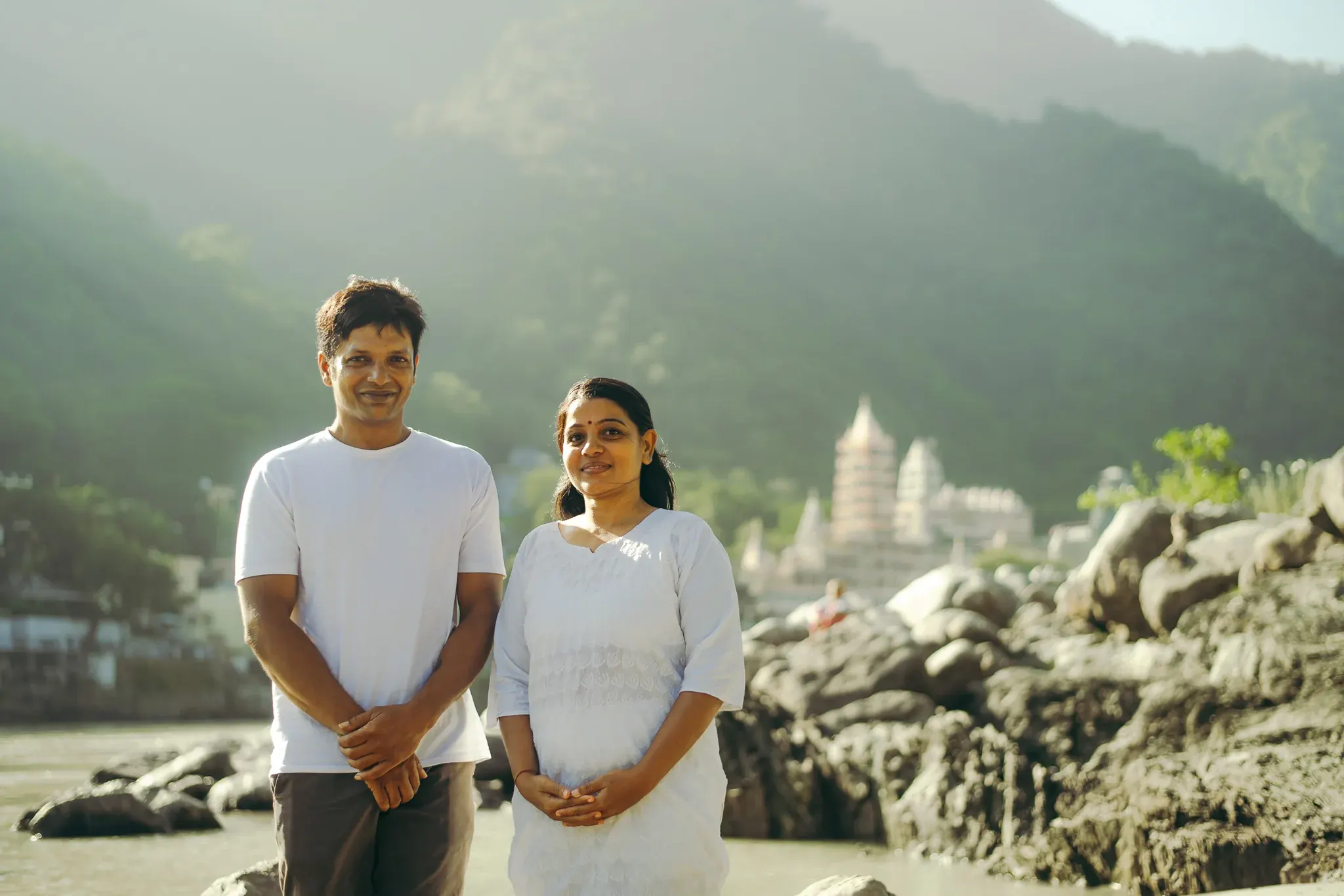 Yoga teacher and a woman in a white kurta smile on the Ganga riverbank in Rishikesh, temple and hills behind