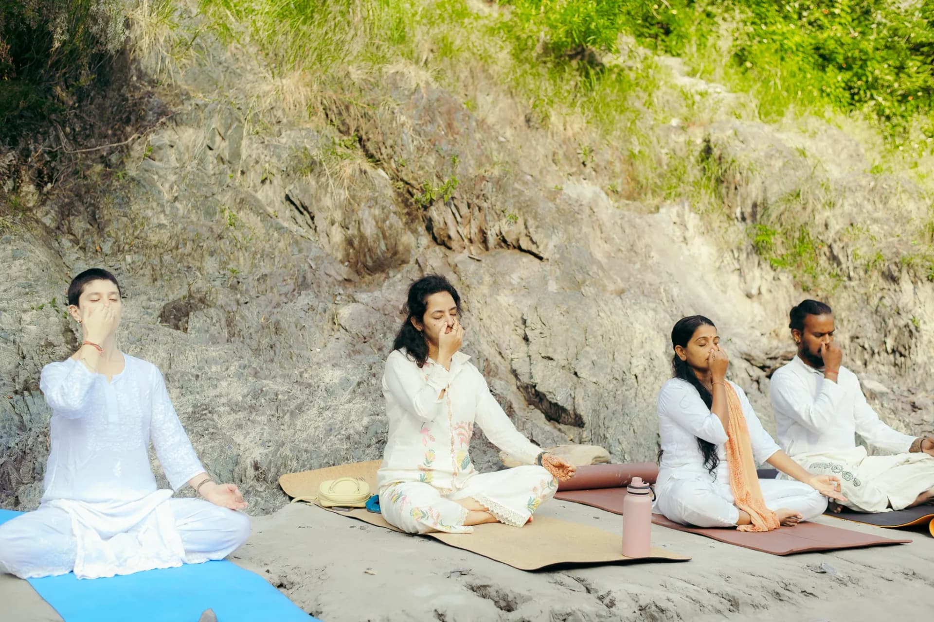 Four students in white practise Nadi Shodhana alternate nostril breathing together on the riverbank in Rishikesh