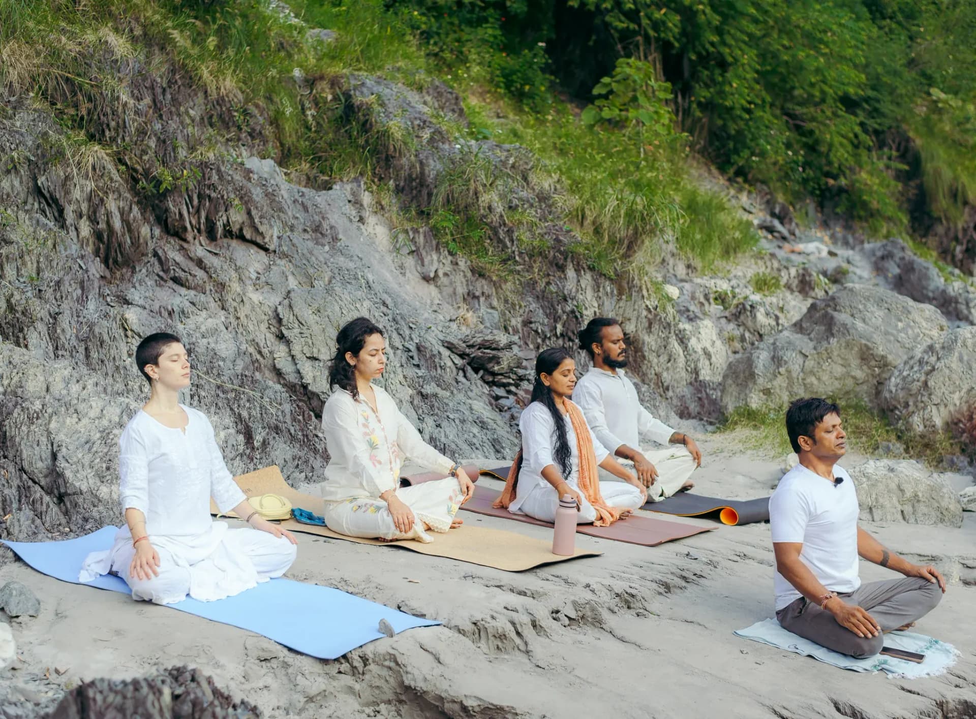 Teacher and four students sit upright with eyes closed on the rocky Ganga riverbank in Rishikesh