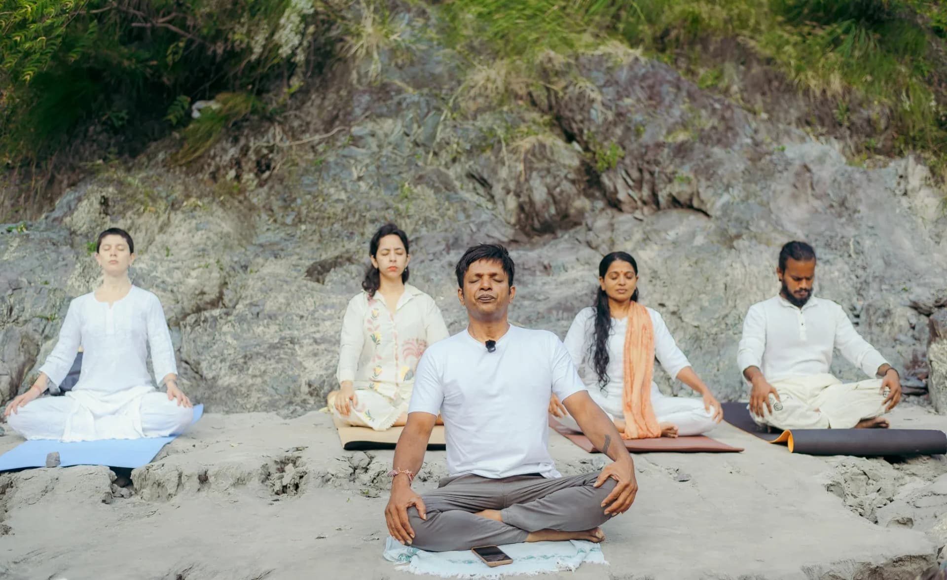 Yoga teacher leads four students in white in seated meditation on the rocky Ganga riverbank in Rishikesh