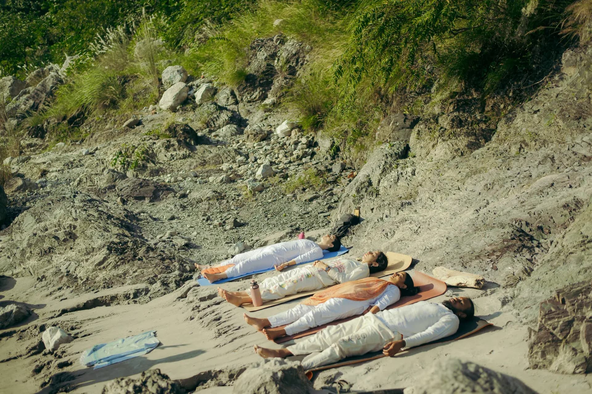 Four students in white rest in savasana on yoga mats on the rocky riverbank in Rishikesh