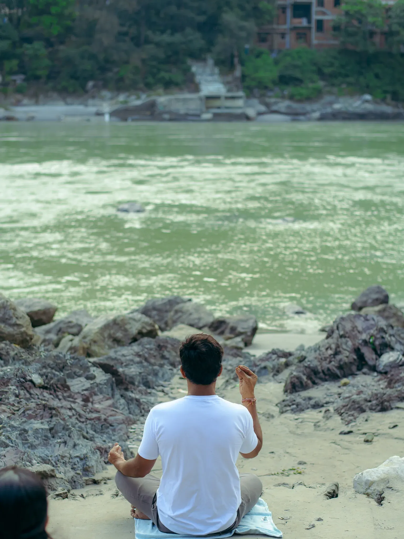 Teacher sits cross-legged on the Ganga riverbank in Rishikesh facing the river, one hand raised in a mudra