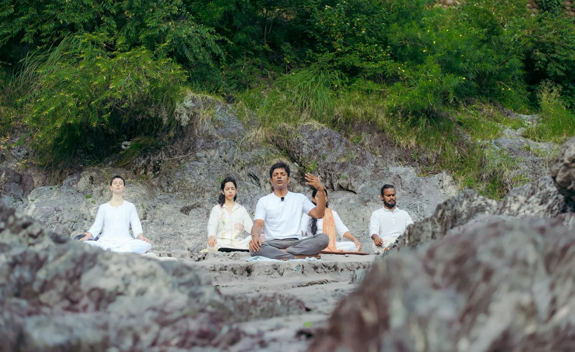 Teacher guides four students in seated meditation on the rocky Ganga riverbank in Rishikesh