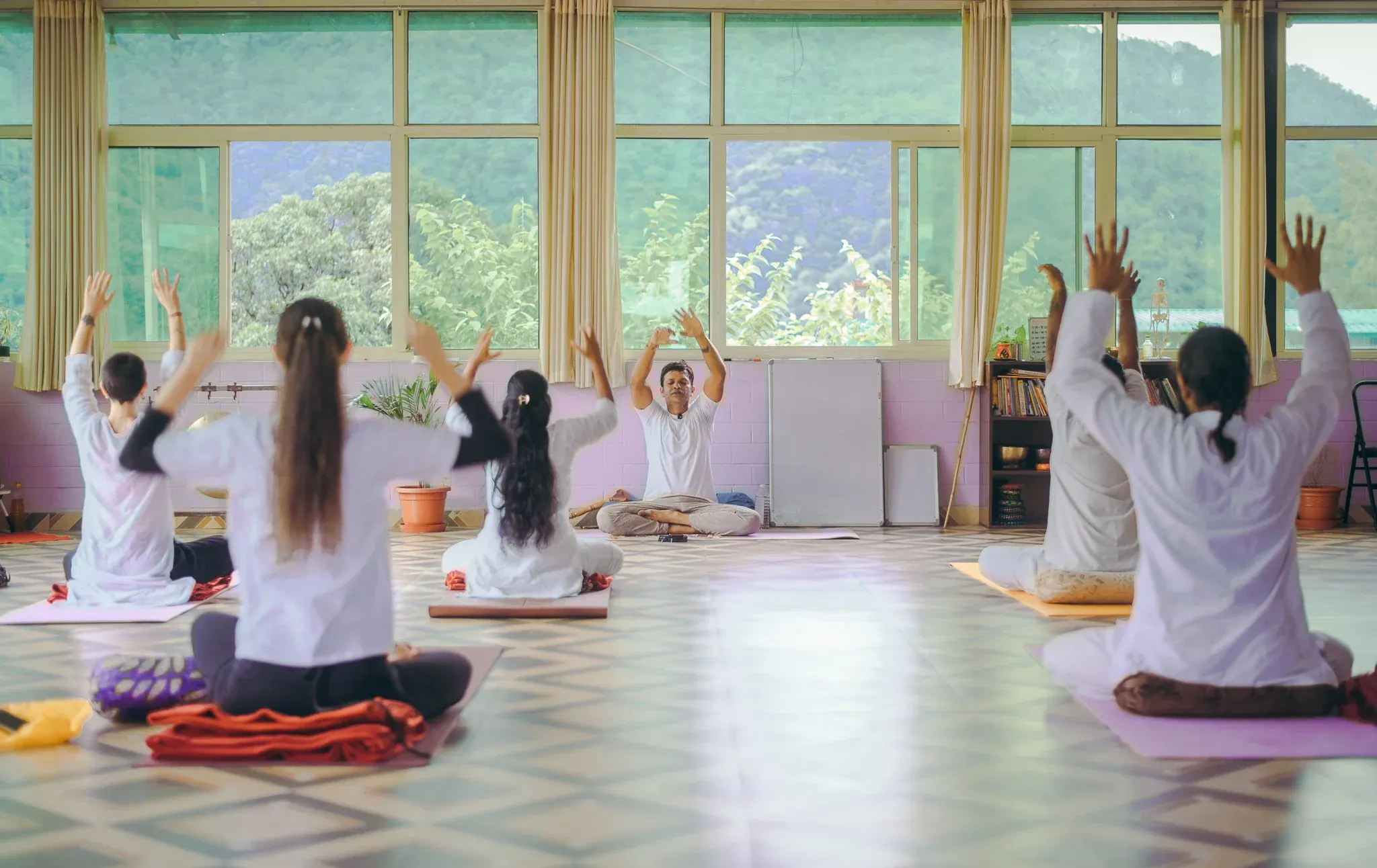 Teacher and students in white raising their arms overhead in a bright yoga shala in Rishikesh