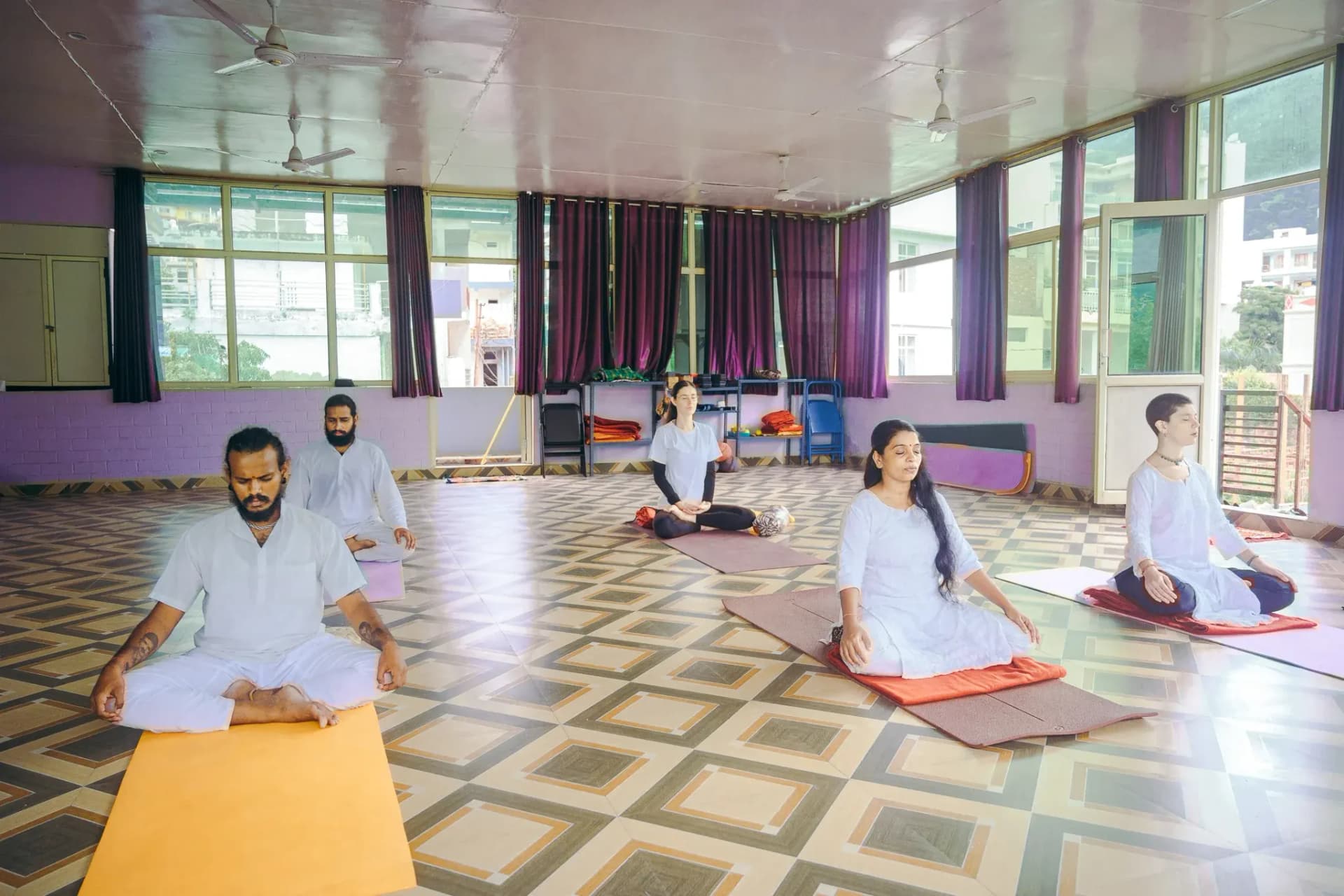 Five students in white meditating cross-legged with eyes closed in a yoga hall in Rishikesh