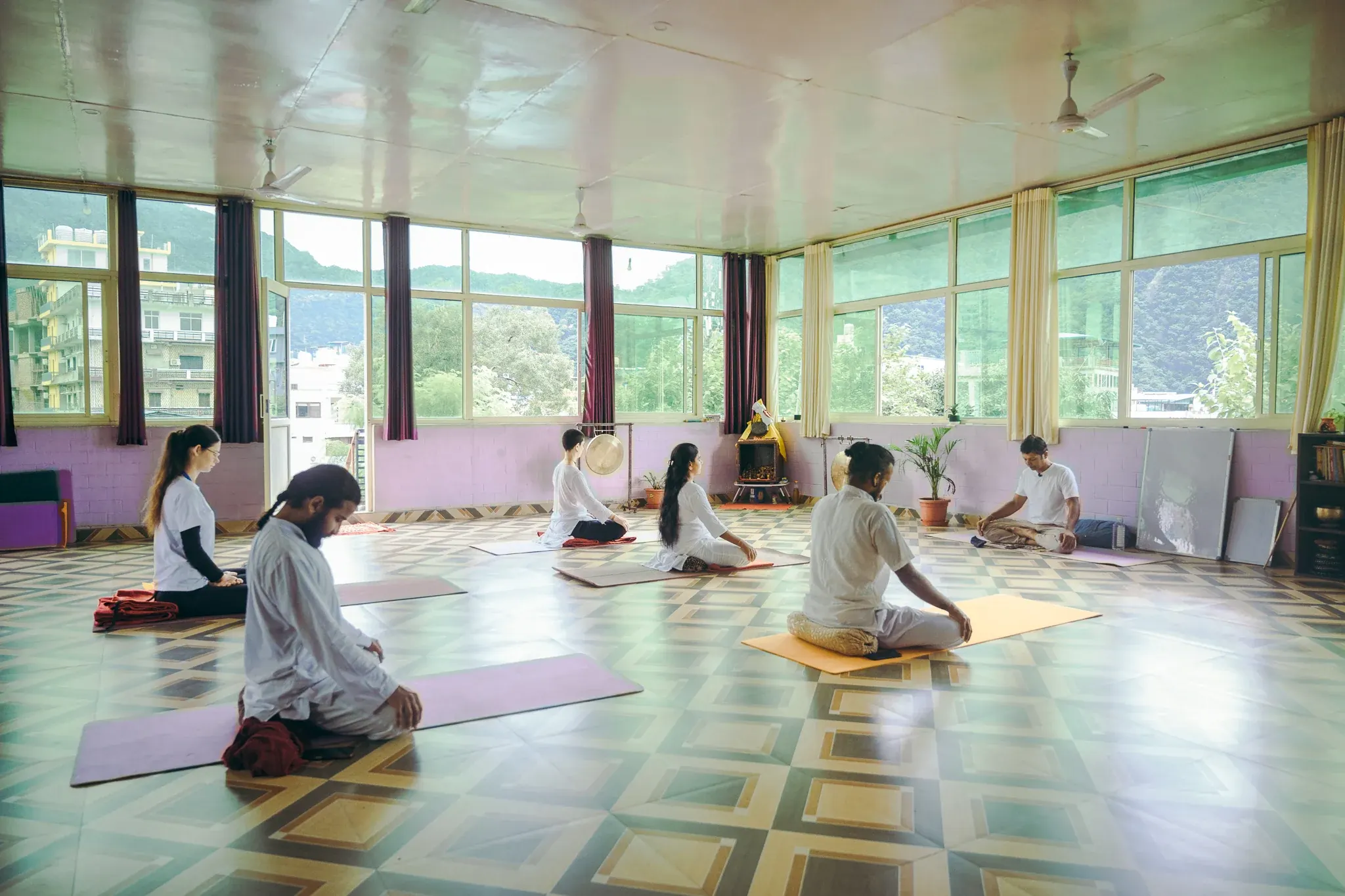 Students in white sit with chins lowered as the teacher guides a class in a large Rishikesh yoga hall