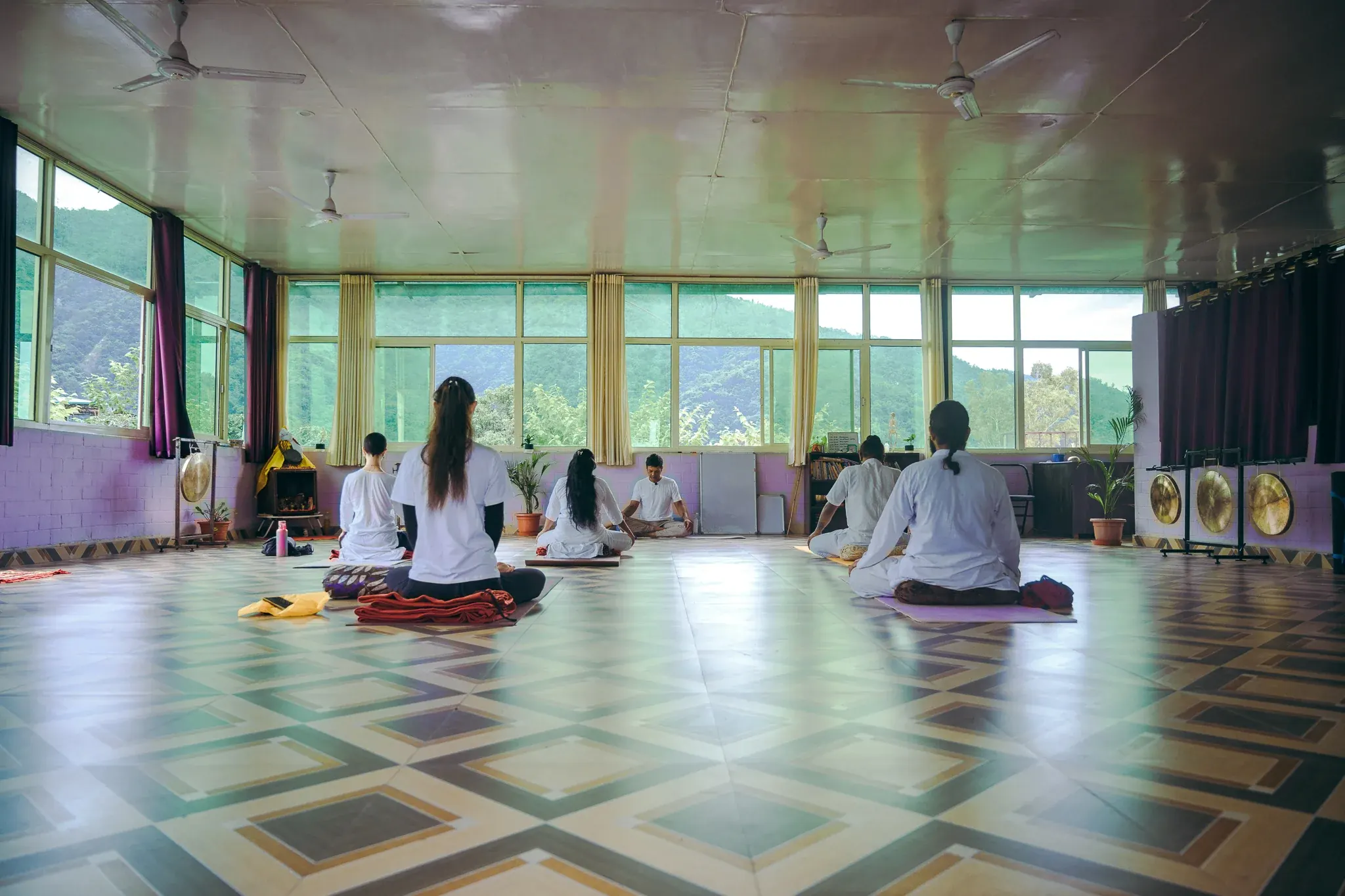 Students sit facing their teacher in a spacious Rishikesh yoga hall with brass gongs and forested hills