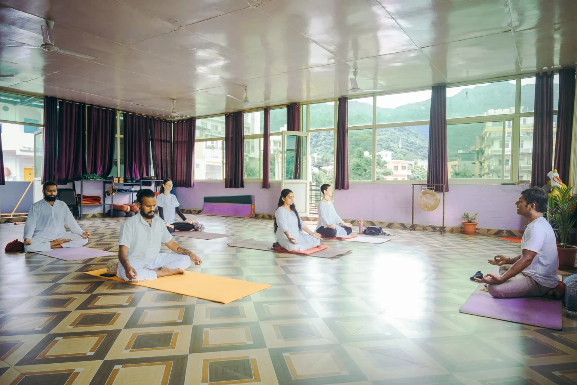 Teacher guides students seated in meditation in a Rishikesh yoga hall with forested hill views