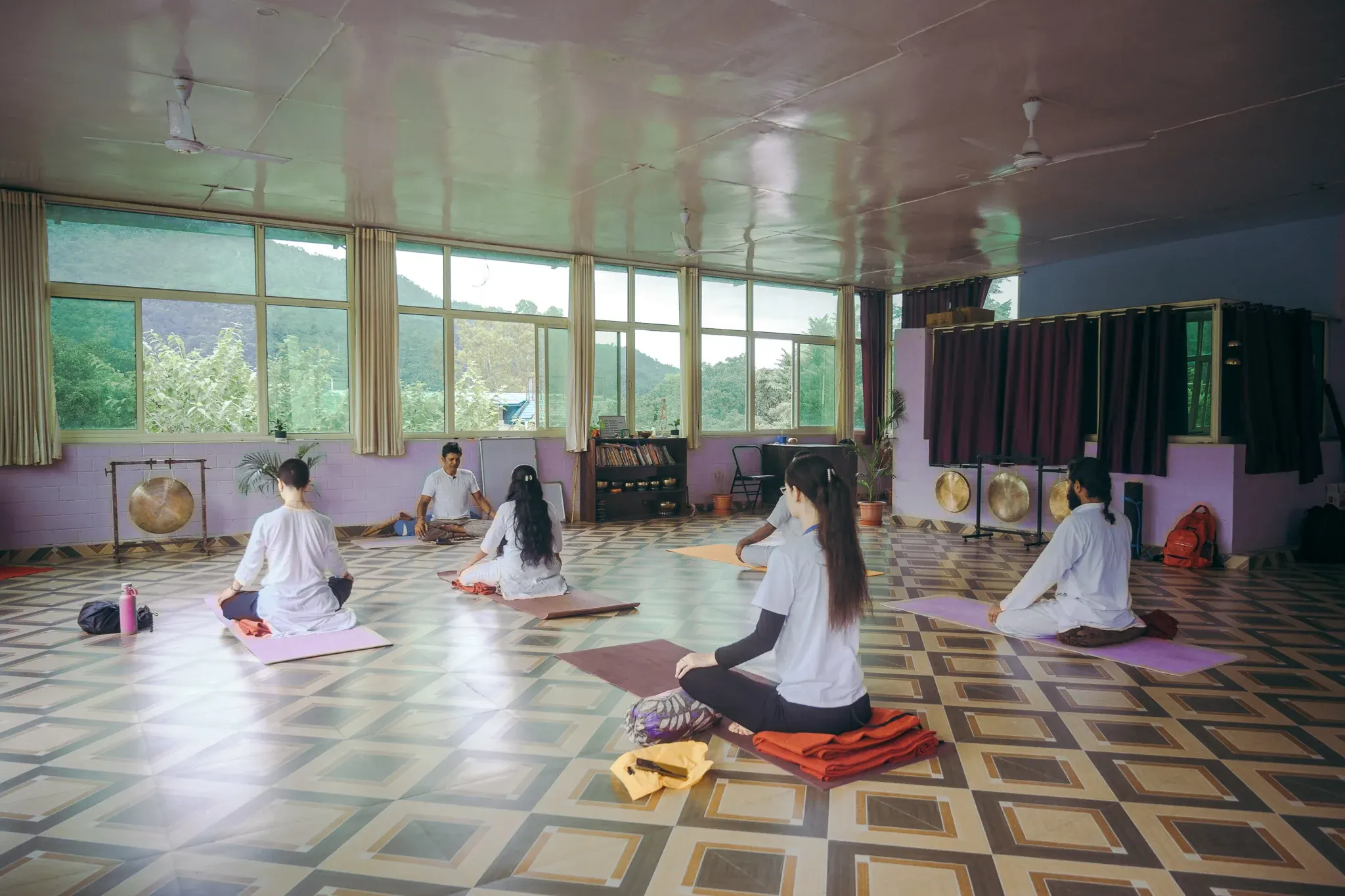 Wide view of a yoga shala in Rishikesh with a teacher and five students seated before windows facing green hills