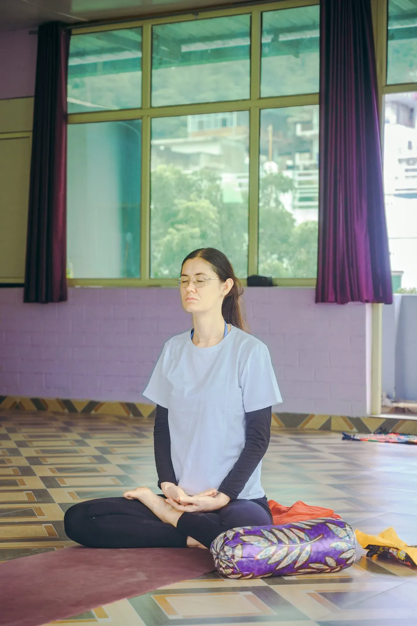 Woman with glasses meditating cross-legged with hands resting in her lap in a yoga shala in Rishikesh