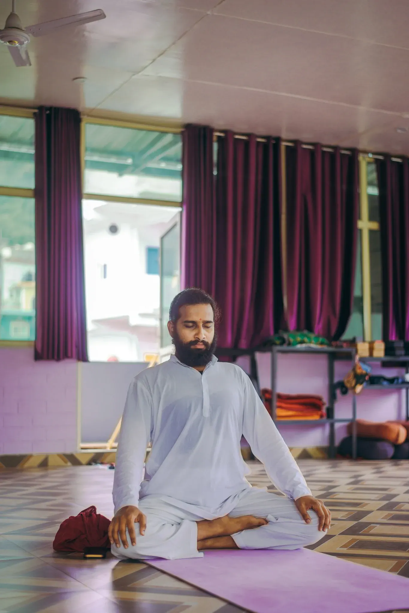 Bearded student in a white kurta sitting cross-legged with eyes closed in a Rishikesh yoga hall