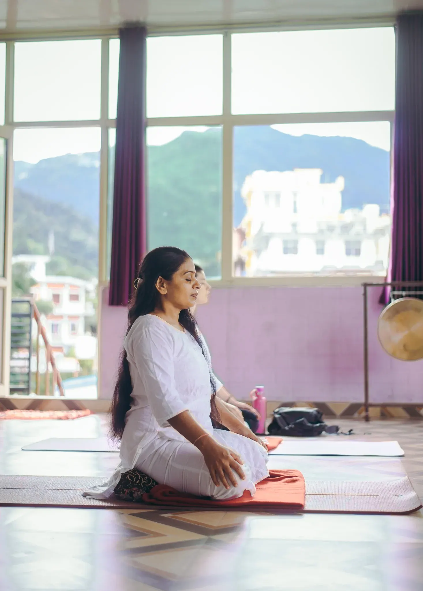 Woman in white meditating in profile in a Rishikesh shala, mountain and town buildings visible through the window