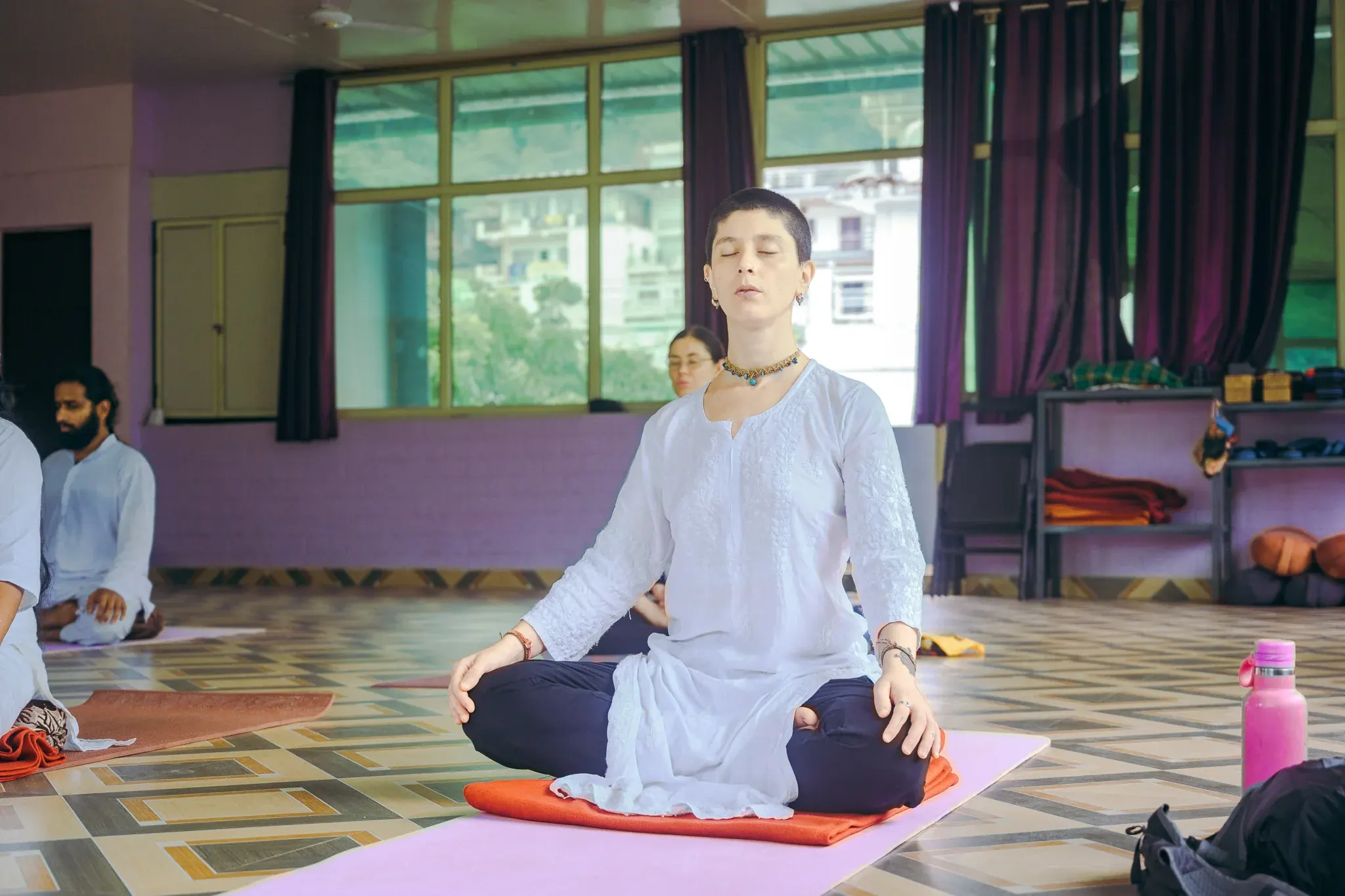 Woman in a white kurta sitting cross-legged with eyes closed and chin raised during practice in a Rishikesh shala