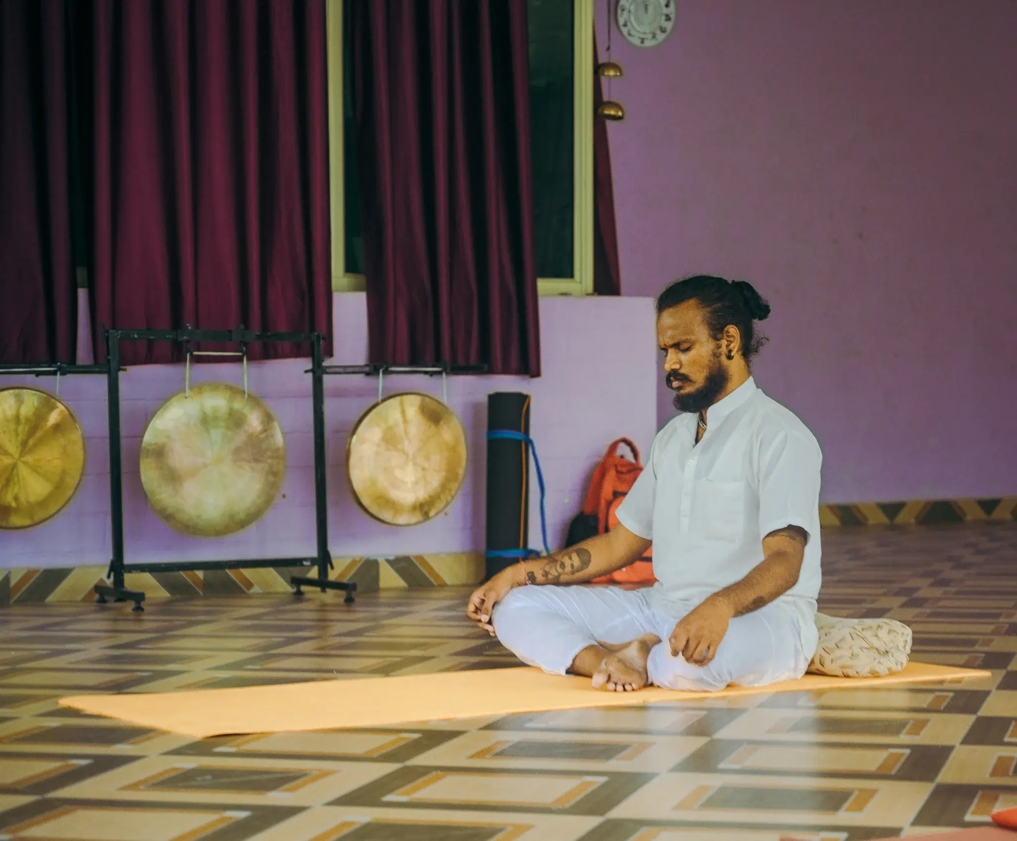 Bearded student in white meditating cross-legged on an orange mat beside three brass gongs in a Rishikesh shala