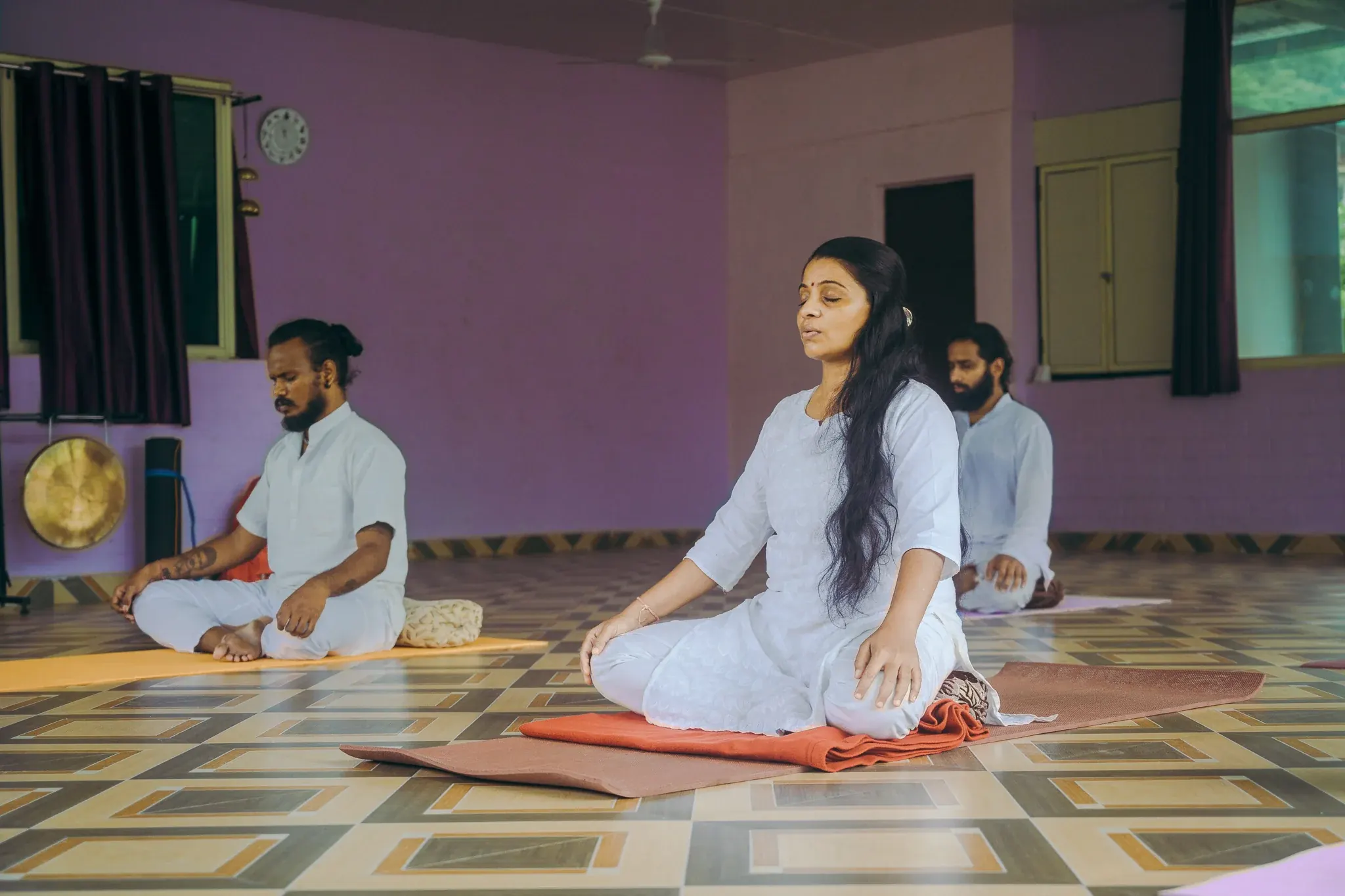 Woman in white sitting cross-legged with eyes closed and lips parted, two men meditating behind her in a Rishikesh shala
