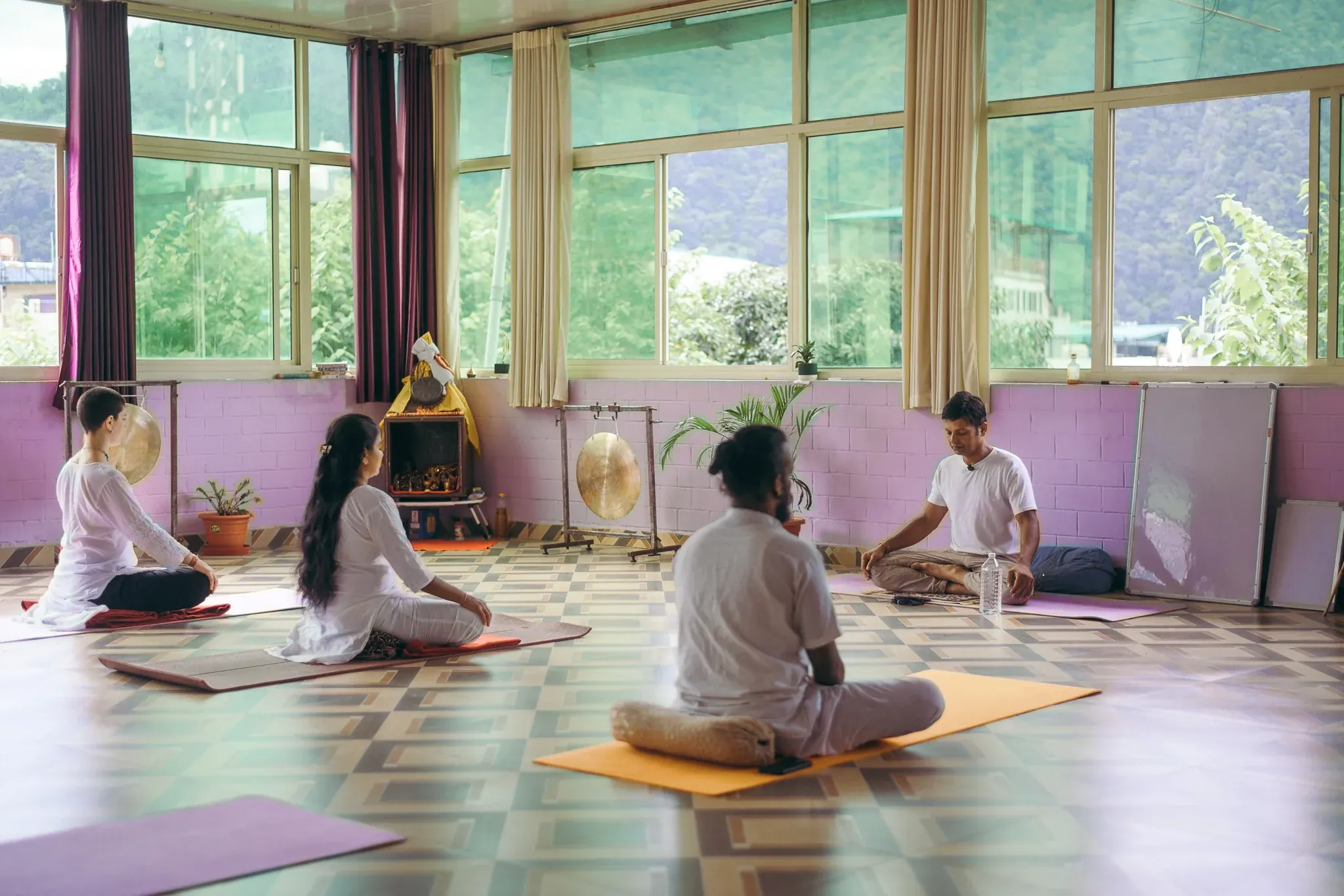 Pranayama teacher leading three students in white in a Rishikesh yoga shala with large windows facing green hills