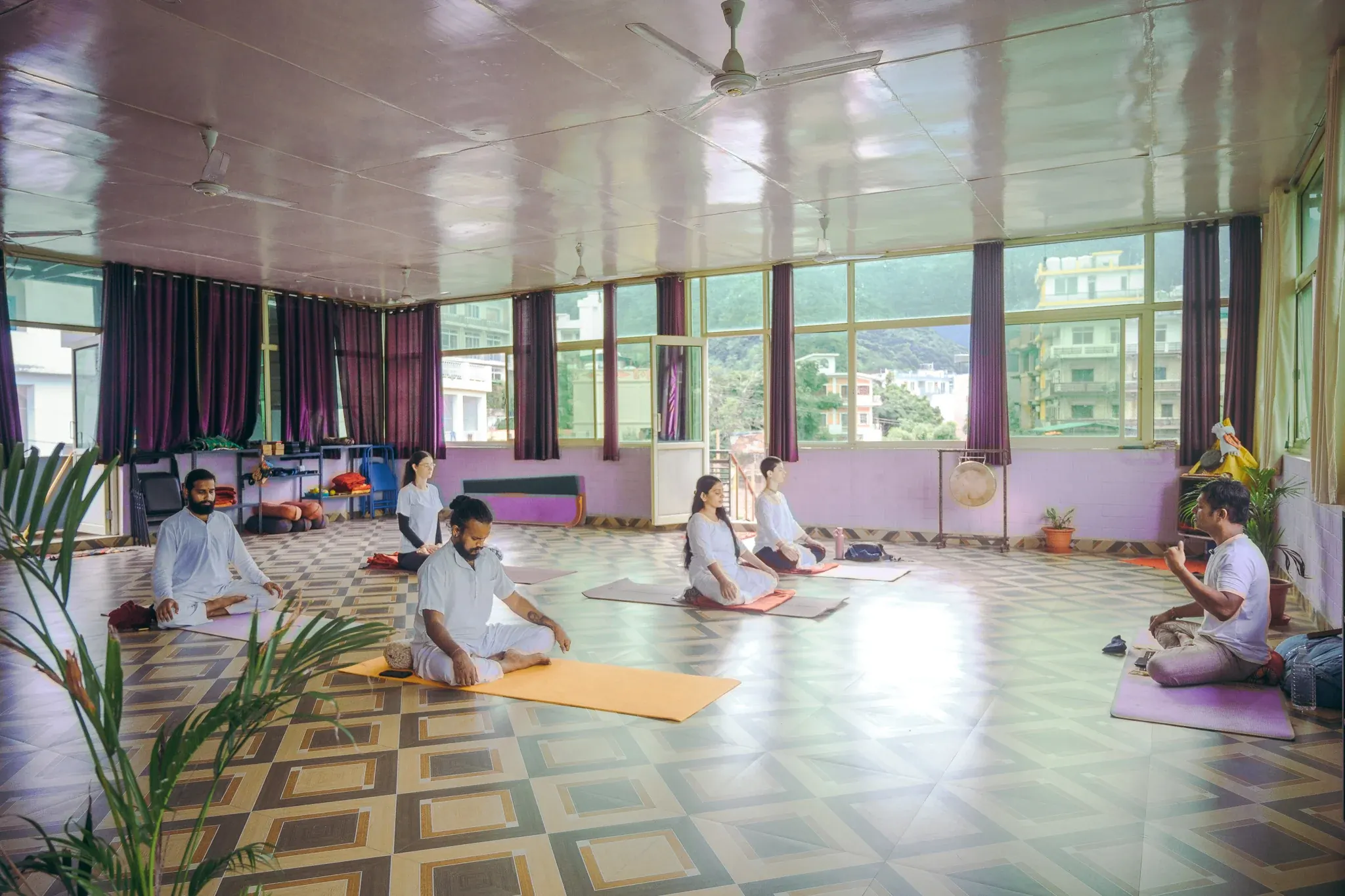 Teacher instructing five students seated in meditation in a spacious Rishikesh yoga hall with views of the hills