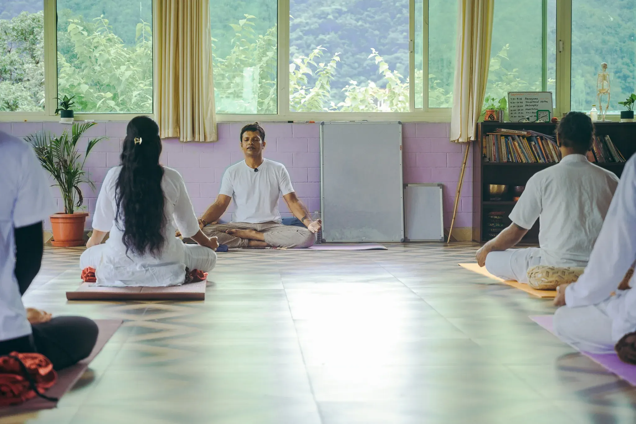 Pranayama teacher sits facing his students in a Rishikesh yoga hall with a bookshelf and hill views