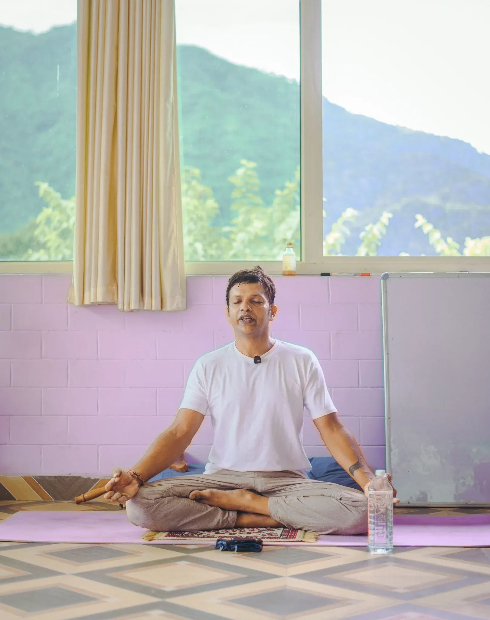 Pranayama teacher seated cross-legged with eyes closed before a window overlooking forested hills in Rishikesh