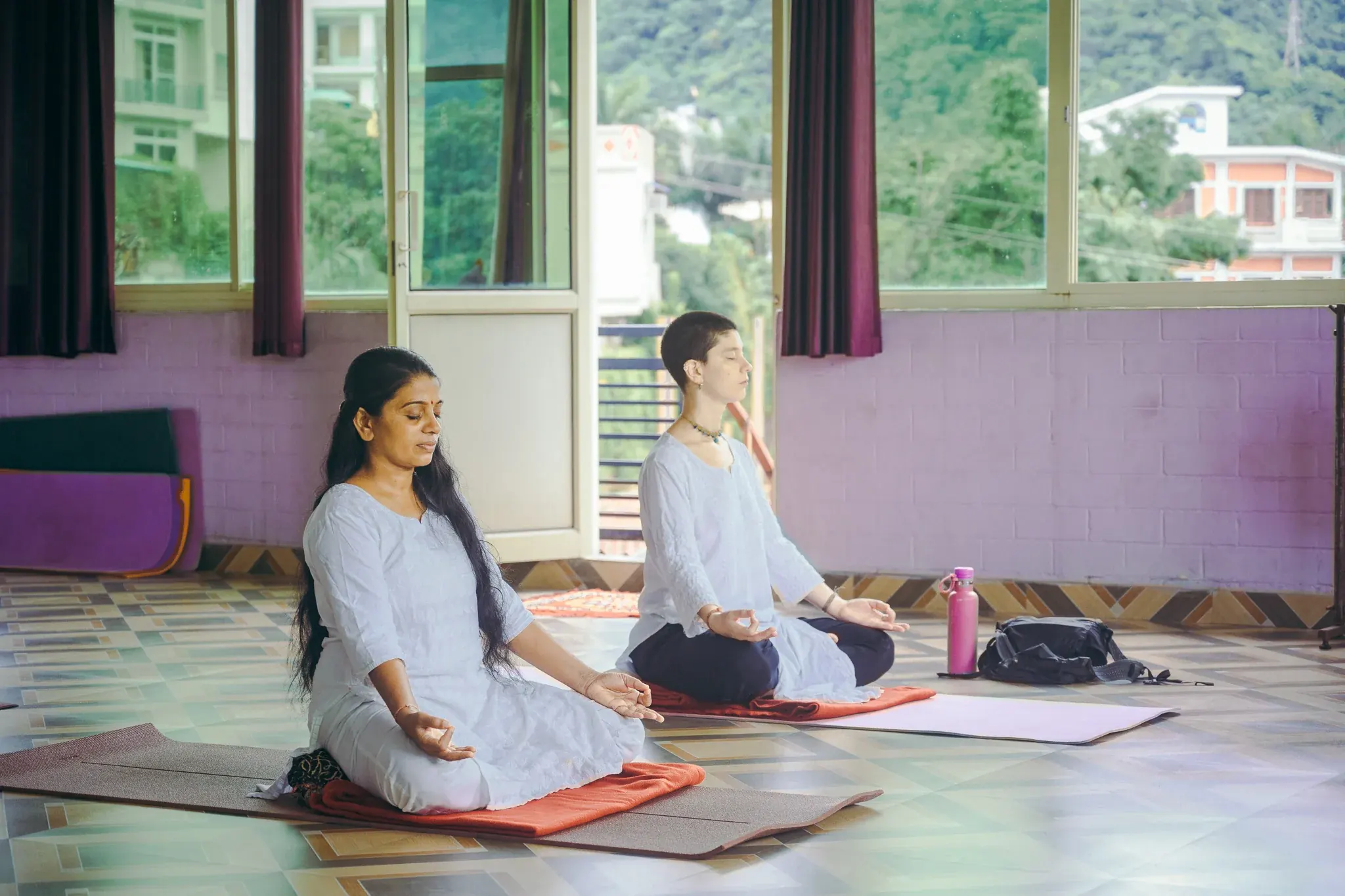 Two women in white meditate with chin mudra on folded orange blankets in a Rishikesh yoga shala